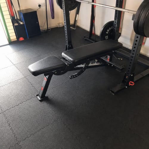 Customer photo: rubber gym tiles under a weight bench and squat rack in a PT studio