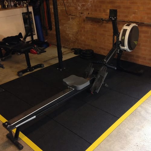 Customer photo: black rubber gym mats with yellow ramp edges under a rowing machine in a home gym